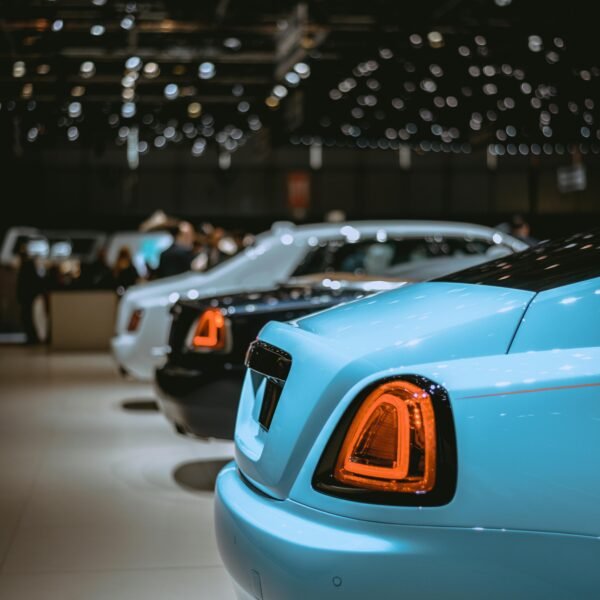 Rear view of luxury cars lined up at an indoor auto show with bright lighting.